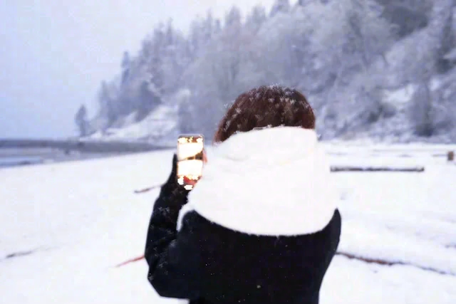 A person seen from behind wears a white scarf and holds a phone up toward a snowy landscape.