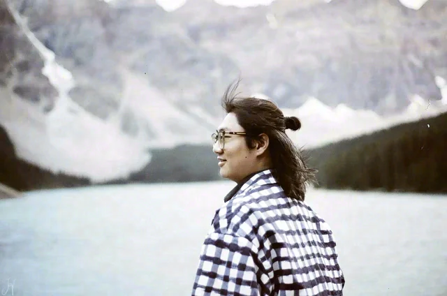 A person wearing glasses and a checkered shirt stands in front of a pale blue lake and mountain slopes.