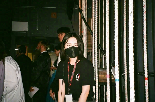A person in a black mask stands beside hanging white ropes, with other people in the dim background.