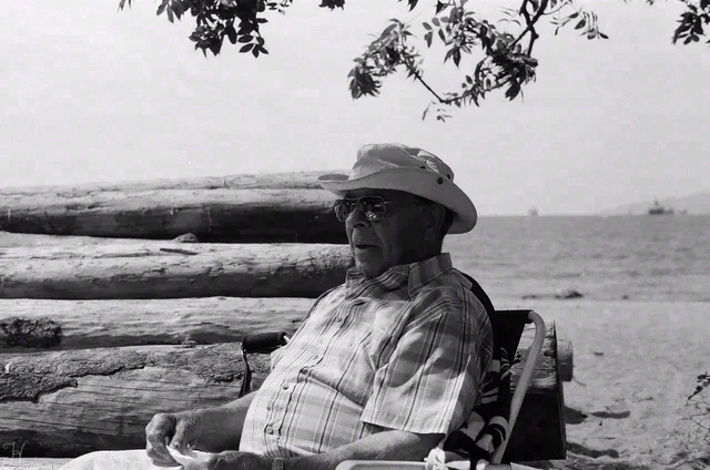 Black and white portrait of a seated person in a sun hat and sunglasses, with driftwood and water behind them.