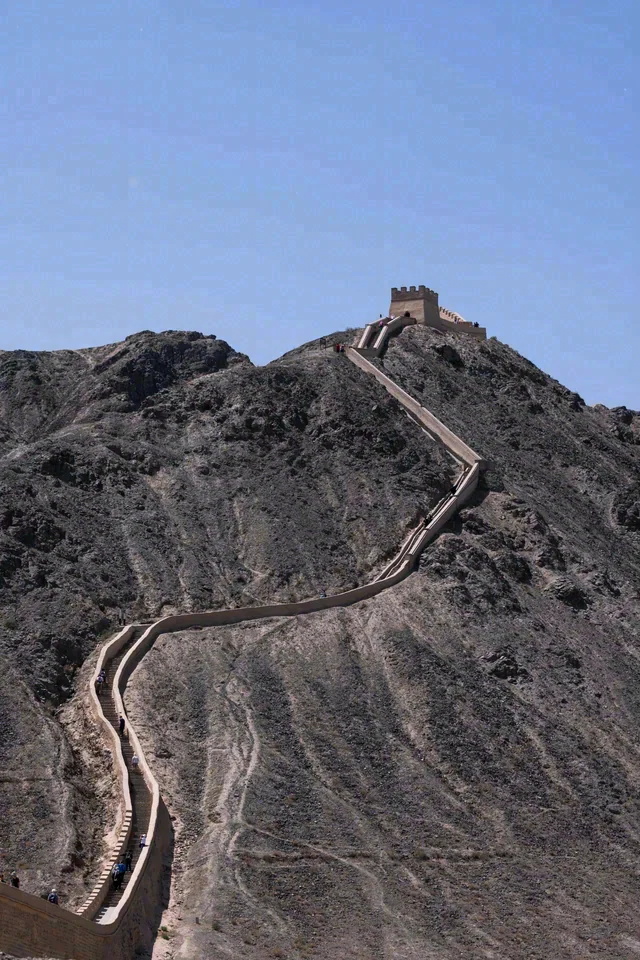 A winding stone wall leads up a barren rocky ridge to a small structure beneath blue sky.
