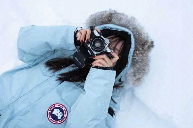 A person in a pale blue hooded coat holds a camera to their face while lying in the snow.