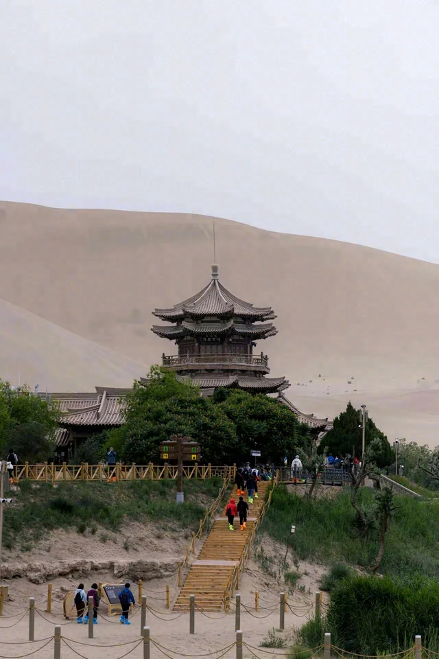 A multi-tiered building stands among green trees beneath a large sand dune, with people in the foreground.