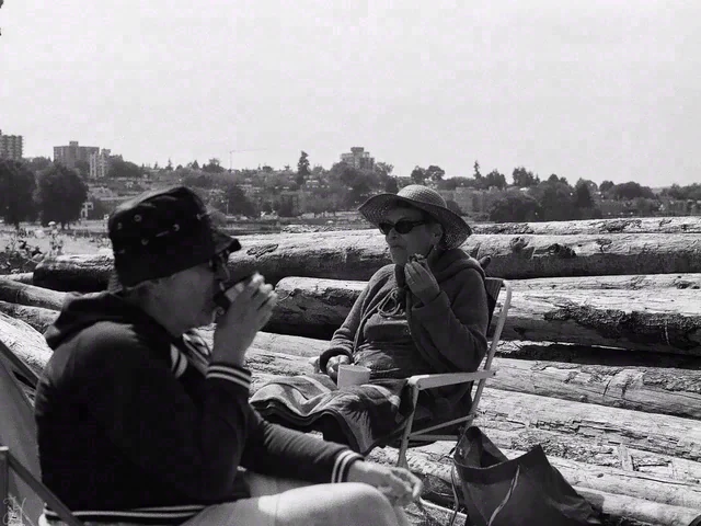 Black and white view of two people seated beside driftwood, with one lifting a cup toward their face.