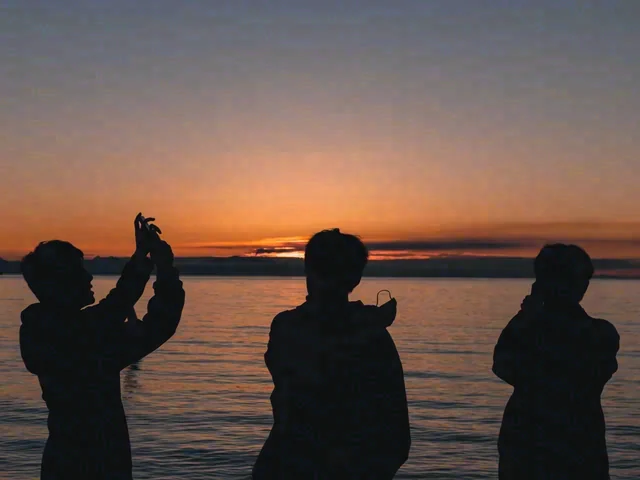 Three people are silhouetted beside the water under an orange evening sky; one holds a phone above their head.